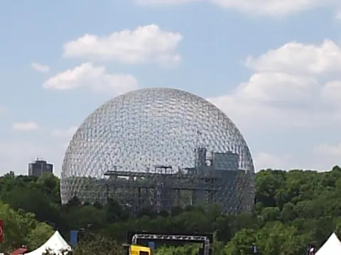 Blick über die Strecke in Montreal auf die geodätische Kuppel von Buckminster Fuller, davor Zuschauer am Zaun und Streckenposten