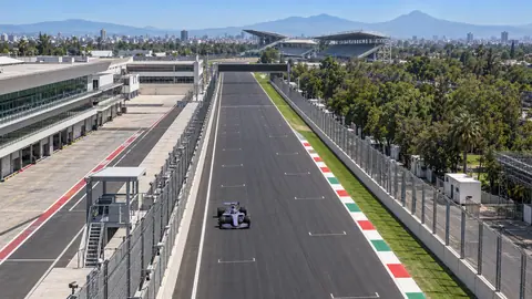 Symbolbild (KI-generiert): Ein Formel-Auto fährt bei Sonne über die Start-Ziel-Gerade des Autódromo Hermanos Rodríguez, links die Boxengasse, hinten Tribünen, Stadt und Berge