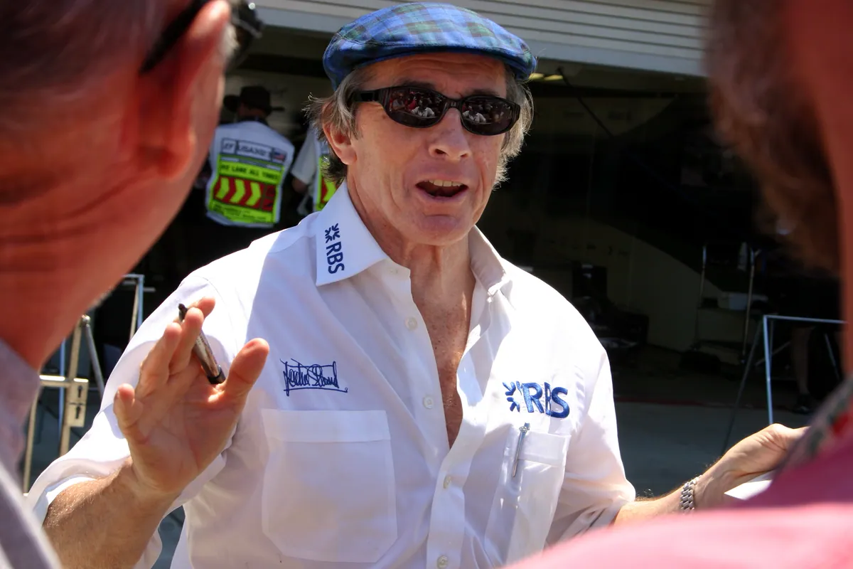Jackie Stewart im weißen Hemd und mit Schirmmütze spricht beim GP USA 2005 in der Boxengasse von Indianapolis mit Fans